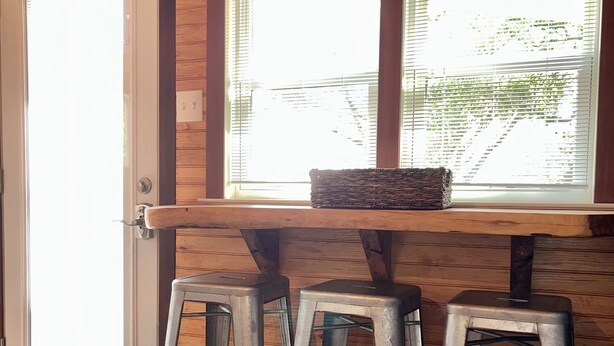 A kitchen with wooden walls, a window with blinds, and three metal stools.