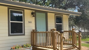 A single-story house with a light green door, a number '365' on the wall, a wooden porch with a bench, and a planter with flowers.