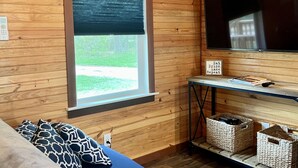 A cozy room with wooden walls, a large window with blinds, a flat-screen TV, a blue sofa, and wicker baskets on a shelf.