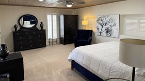 Un dormitorio con techo de madera, una cama con edredón blanco, un sillón azul oscuro, un aparador, un espejo, un cuadro y una ventana con persianas.