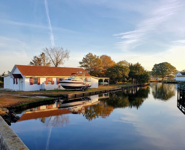 A house with a boat docked outside, a calm water body reflecting the surroundings, and a clear sky. - The Lucky Hook, Hyde Co, Waterfront, Pet & Family Friendly w/kayaks, Fireplace (Engelhard)