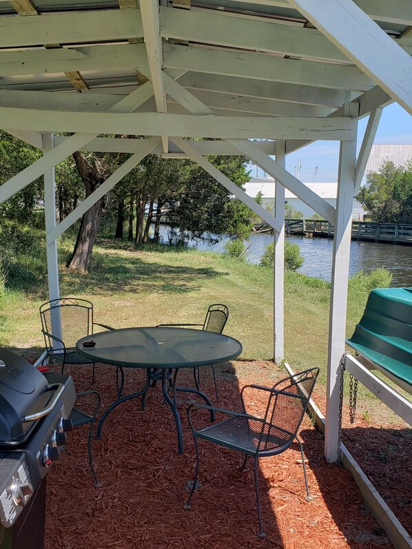 Un patio techado al aire libre con una parrilla, mesa y sillas, con vistas a un cuerpo de agua y follaje.