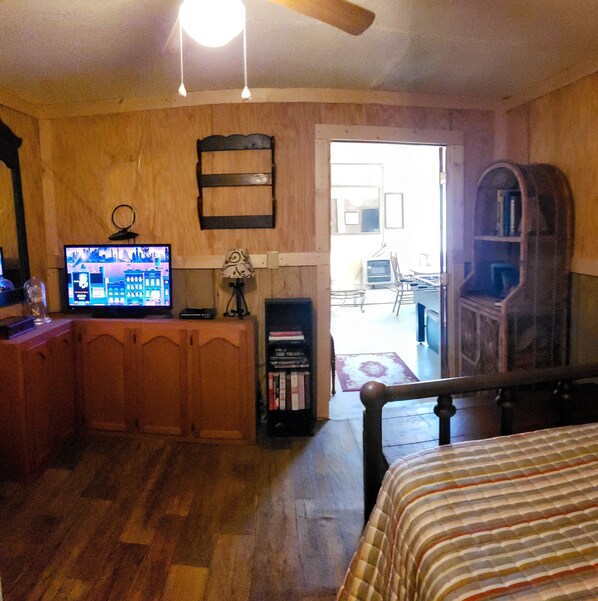 A room with a wooden floor, a bed, a television, a bookshelf, and a door leading to another room.