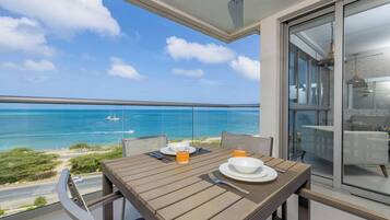 Varanda com mesa posta para dois, vista para uma praia sob um céu azul límpido e oceano.