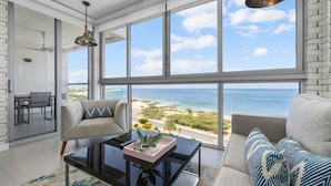 Een moderne woonkamer met een groot raam met uitzicht op een strand, een bank, een bijzettafeltje en een hanglamp.