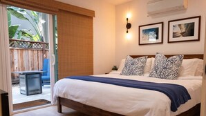 Una camera da letto con un letto di legno, biancheria bianca, cuscini decorati, una coperta blu, una porta vetrata scorrevole e una piccola pianta sul comodino.