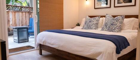 A bedroom with a wooden bed, white bedding, patterned pillows, a blue throw, a sliding glass door, and a small plant on the bedside table.