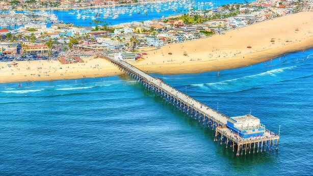Un pontile si protende verso il mare, con una barca ormeggiata in fondo, circondato da una spiaggia e una cittadina costiera.