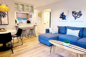 A modern living room with a blue sofa, a dining table with chairs, a kitchen area with a countertop, and a hanging light fixture.