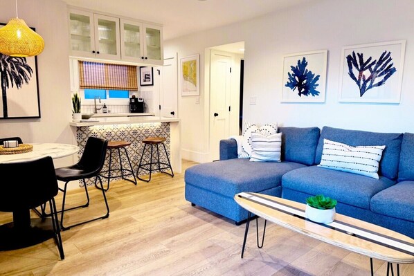 A modern living room with a blue sofa, a dining table with chairs, a kitchen area with a countertop, and a hanging light fixture.