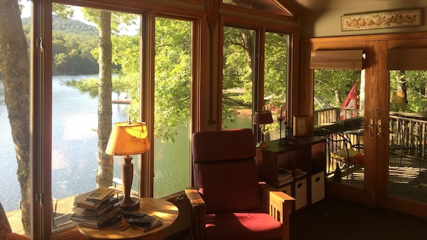 Un chalet confortable avec de grandes fenêtres donnant sur un lac, un fauteuil rouge et une table en bois sur laquelle sont posés des livres et une lampe.
