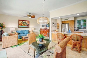 A modern living room with a glass coffee table, wicker furniture, a ceiling fan, and a kitchen area with a refrigerator and counter.