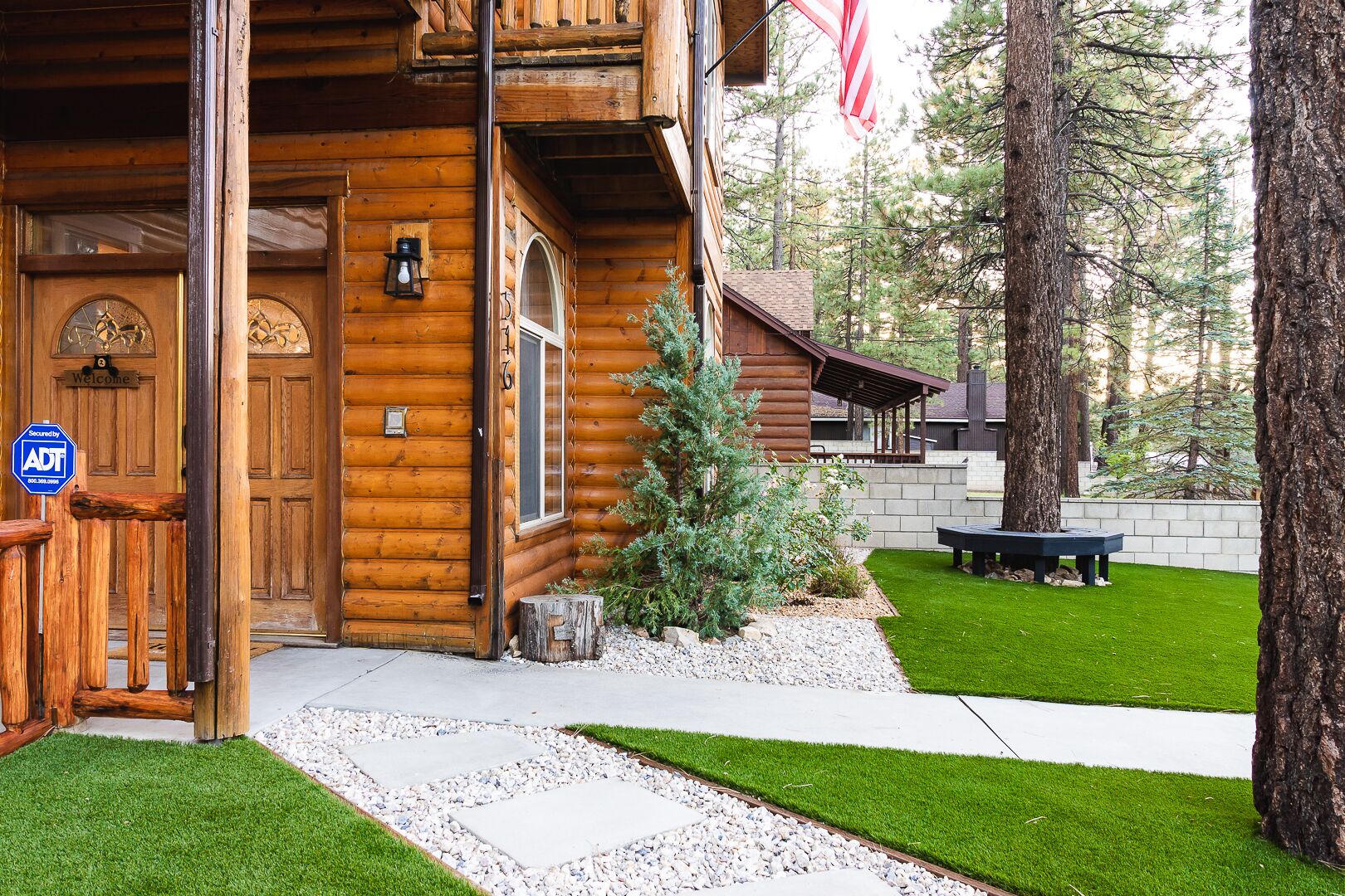 A log cabin with a wooden door, a welcome sign, a small porch, and a flag.
