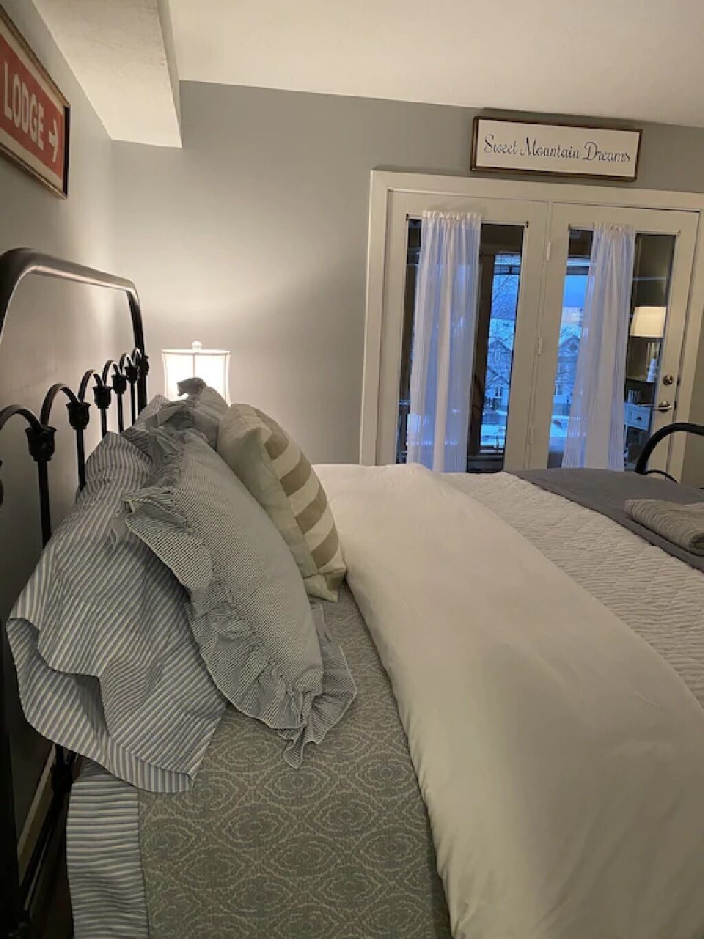 En pænt redt seng med stribede og ensfarvede puder, en sengegavl med et bøjet design, og et skilt over døren med teksten "Sweet Mountain Dreams".