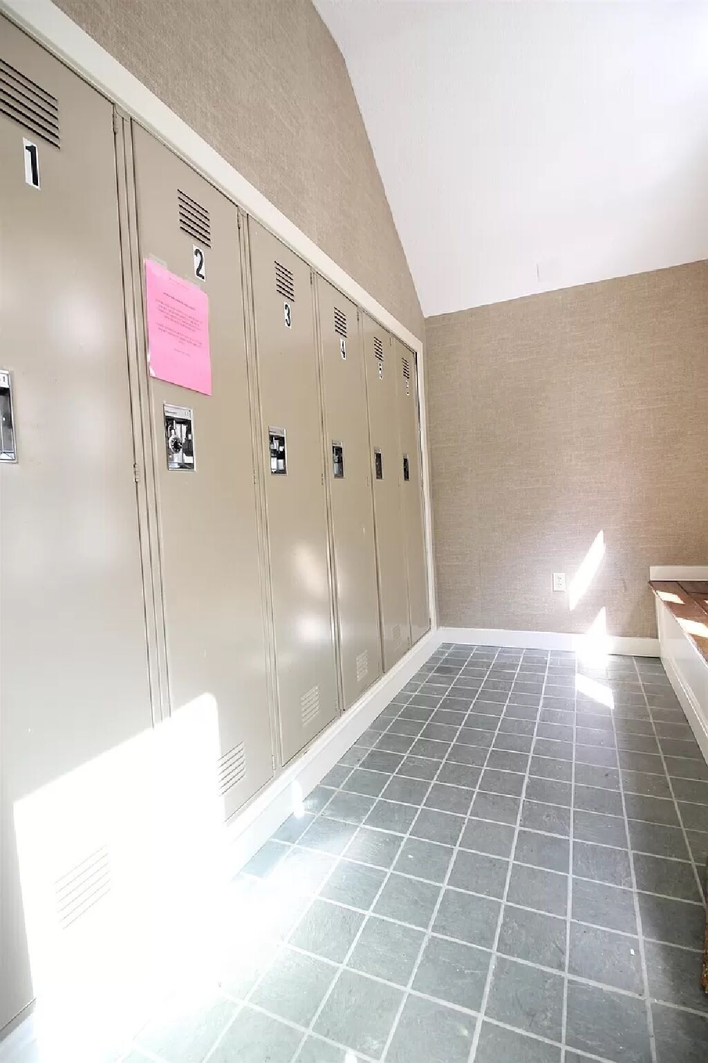 Un vestiaire avec des casiers beiges numérotés de 1 à 3, un avis rose et un sol carrelé.