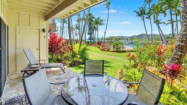 A covered patio with a glass table and chairs, overlooking a well-manicured lawn with palm trees and a view of the ocean. - Lae Nani 114.  Beachfront, Ground Floor Condo (Kapaʻa)
