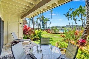 A covered patio with a glass table and chairs, overlooking a well-manicured lawn with palm trees and a view of the ocean. - Lae Nani 114.  Beachfront, Ground Floor Condo (Kapaʻa)