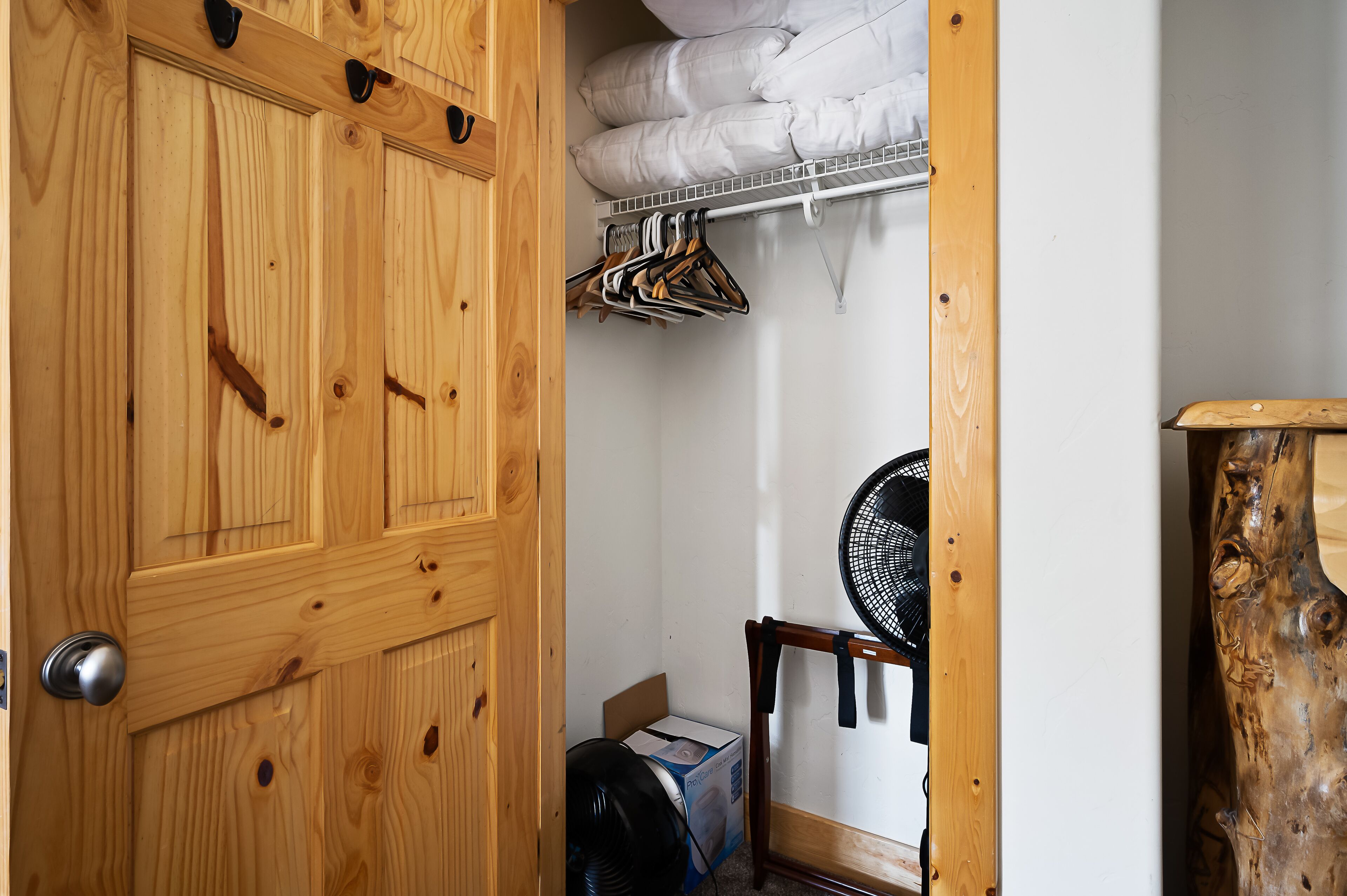 Une porte en bois avec une poignée, un placard rempli de vêtements, un ventilateur et une boîte posée sur le sol.