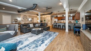 A spacious living room with a large sectional sofa, a coffee table, and a patterned rug. There is a kitchen area with wooden cabinets and a bar counter.