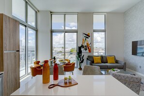 Ett modernt vardagsrum med ett stort fönster, en grå soffa med gula kuddar och en matplats med orange stolar och ett vitt bord.