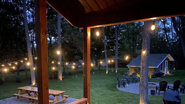 Un jardín con un quiosco de madera, luces colgantes, una mesa de picnic y una zona de barbacoa.