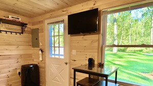 Una cabaña con interior de madera, una puerta con ventana, un televisor sobre la puerta, una mesita y una silla.