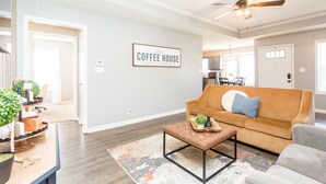 A living room with a brown sofa, a coffee-themed wall sign, and a ceiling fan. - Once-In-A-Lifetime Coffee-Themed Stay For Up To 8. (Clarksville)