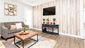 A living room with a grey sofa, a brown ottoman, a wooden console table with plants, and a flat-screen TV mounted on a wooden wall. - Once-In-A-Lifetime Coffee-Themed Stay For Up To 8. (Clarksville)