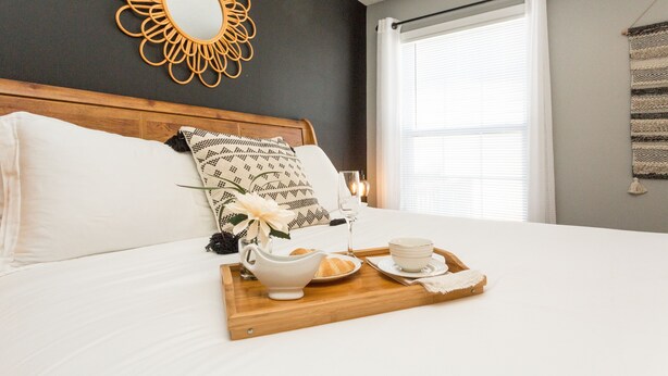 A neatly made bed with a tray of breakfast items, a decorative mirror, and a window with curtains. - Once-In-A-Lifetime Coffee-Themed Stay For Up To 8. (Clarksville)