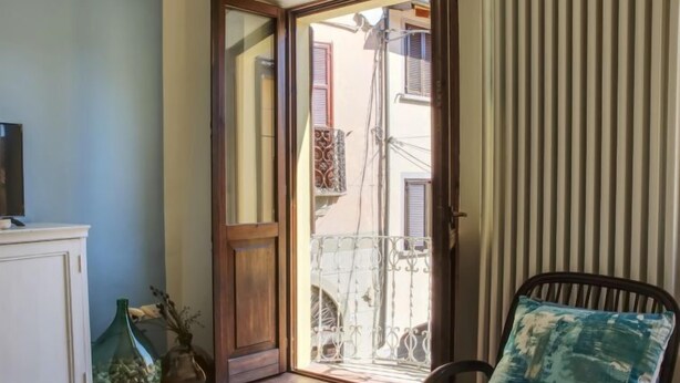 A room with a sliding glass door leading to a balcony, a white cabinet, a green vase, and a chair with a blue cushion. - Canton Balin by Interhome (Meina)