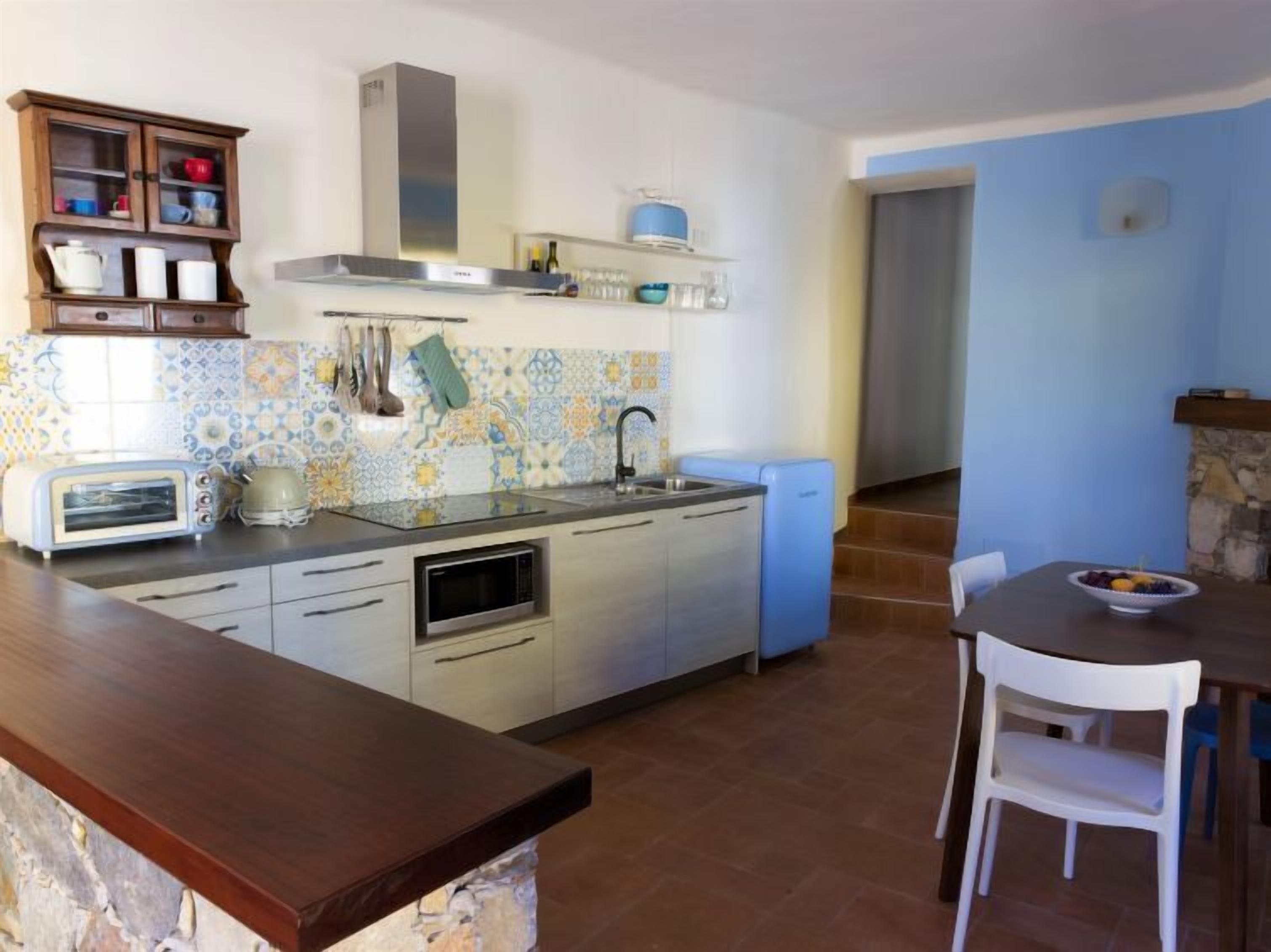 Een kleine keuken met een ingebouwde magnetron, een broodrooster en een spoelbak. Er is een eethoek met een tafel en stoelen, en een stenen muur op de achtergrond.