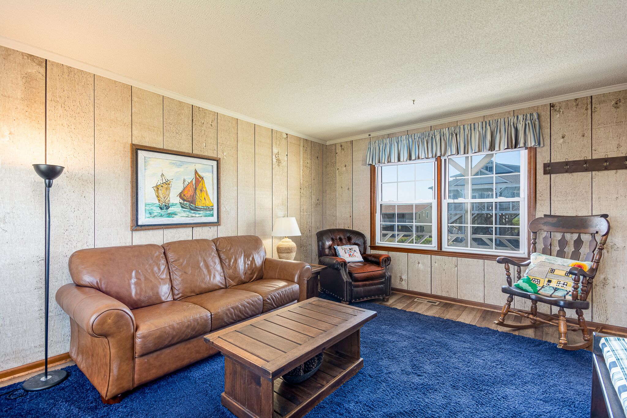 A living room with a brown leather sofa, a wooden coffee table, a rocking chair, and a large window with a view of a house.