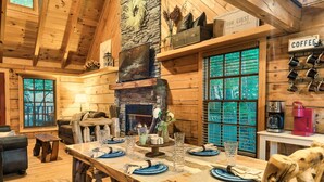 Ein Holzblockhaus-Interieur mit einem Esstisch für vier Personen, einem Kamin aus Stein und einer Kaffeestation.