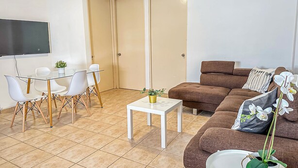 A living room with a brown sofa, a coffee table, and a TV mounted on the wall. There is a dining area with a glass table and white chairs. - Boulevard 107 (Mayagüez)