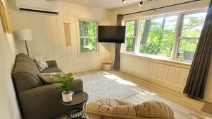 Un salon comprenant un canapé, une petite table avec une plante, un fauteuil en osier, un téléviseur fixé au mur et de grandes fenêtres avec des rideaux.