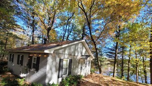 Une petite maison avec un toit en pente, entourée d’arbres et bordée par un plan d’eau.