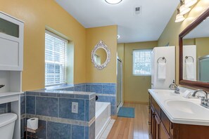 Une salle de bain avec une baignoire, un lavabo et un miroir. Des serviettes sont accrochées au mur et il y a une fenêtre avec des stores.