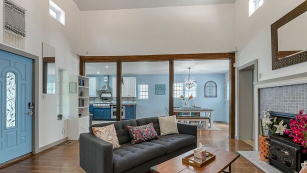 A spacious living room with a dark grey sofa, a wooden coffee table, and a patterned rug. A kitchen with blue cabinets and a dining area with a chandelier are visible in the background. - Norfolk Beach Casita / Fenced Yard / Dog Friendly (Norfolk)