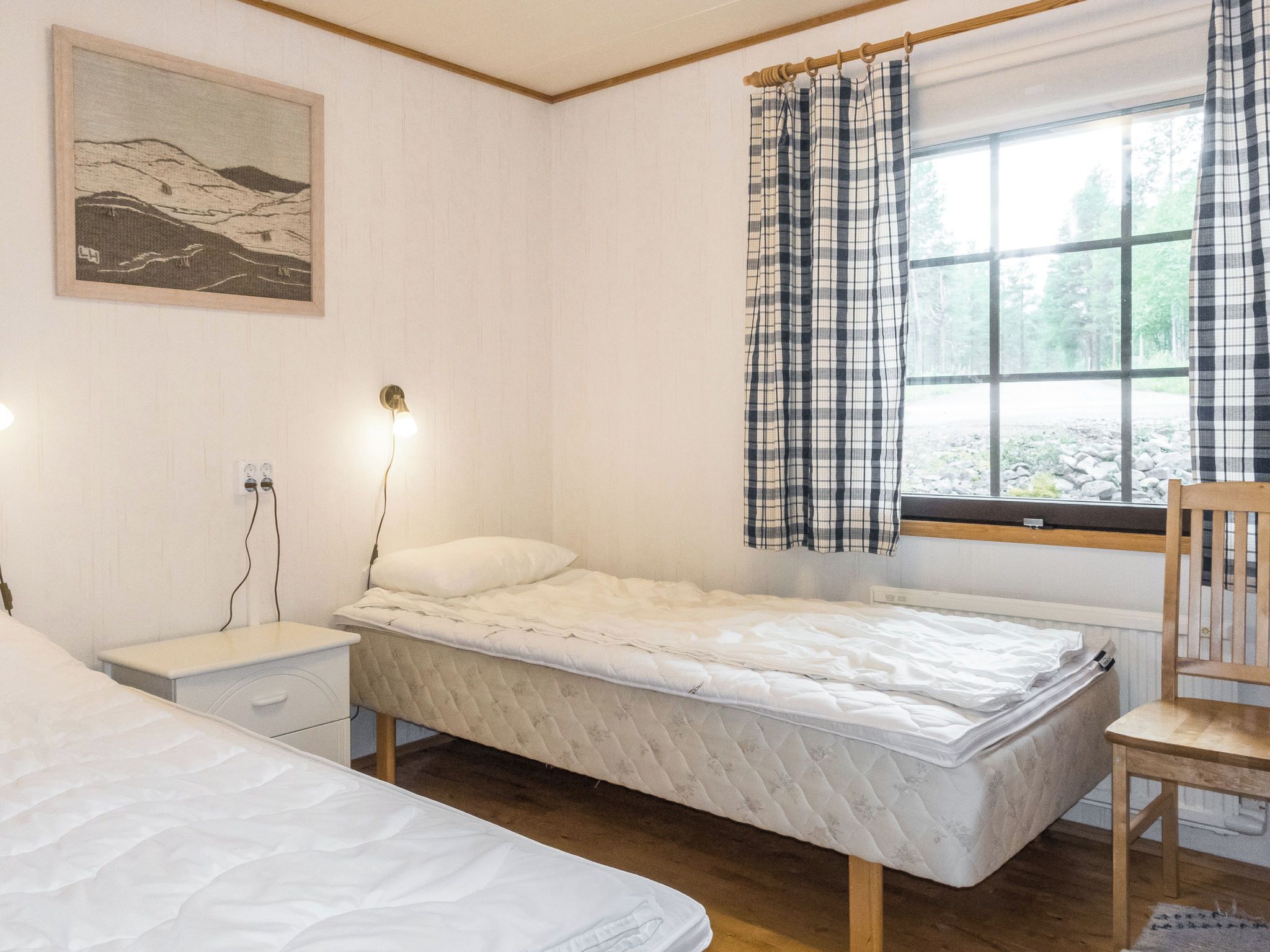 Makuuhuoneessa on kaksi sänkyä, yöpöytä, lamppu, ikkuna, jossa on verhot, ja seinällä on taulu.