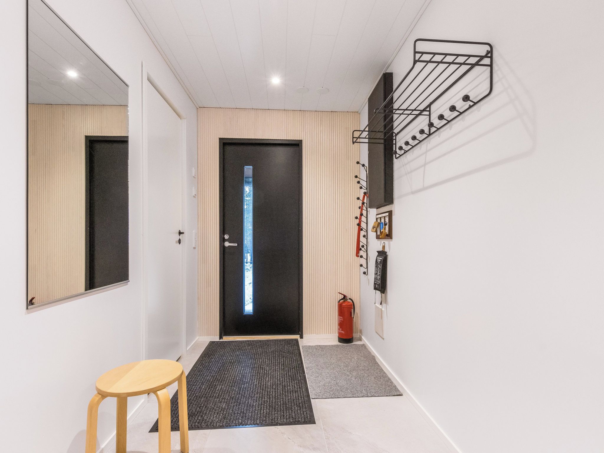 A hallway with a wooden door, a small stool, a fire extinguisher, and a wall-mounted coat rack.