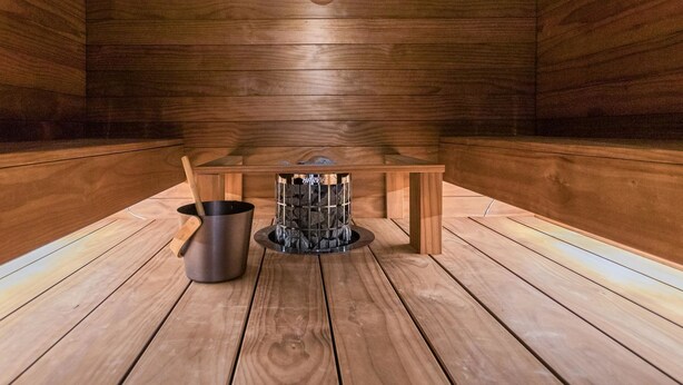 Un sauna avec des murs et un sol en bois, un banc en bois, un appareil de chauffage pour sauna et un seau en métal.