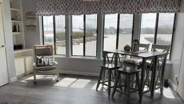 Una habitación con una mesa de comedor y sillas, una ventana que da a una masa de agua, y una cortina estampada.