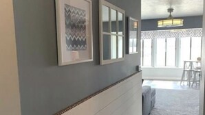 Un pasillo con pared gris, zócalo blanco y alfombra gris. Hay cuadros enmarcados en la pared y una ventana con vista a un patio.