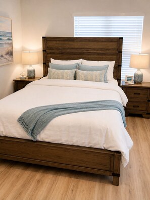 Un dormitorio con cama de madera, ropa de cama blanca, dos mesitas de noche con lámparas, un cuadro en la pared y una ventana con persianas.