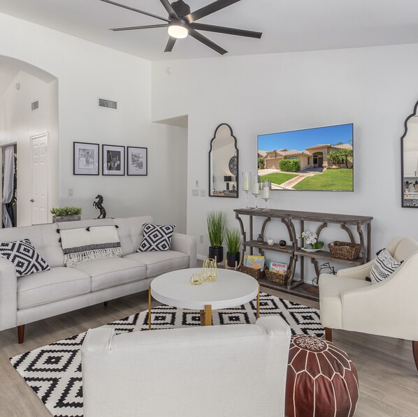 Ein modernes Wohnzimmer mit Sofa, Sessel und rundem Couchtisch. An der Wand befinden sich gerahmte Bilder, ein großer Spiegel und ein Fernseher, der ein Haus zeigt.