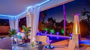 An outdoor dining area with a glass table, wicker chairs, and a pool with illuminated plants. - LAKEFRONT!FREE HEATED POOL,SPA. BOAT, KAYAK. EV charger. Ocotillo Chandler.  (Chandler)
