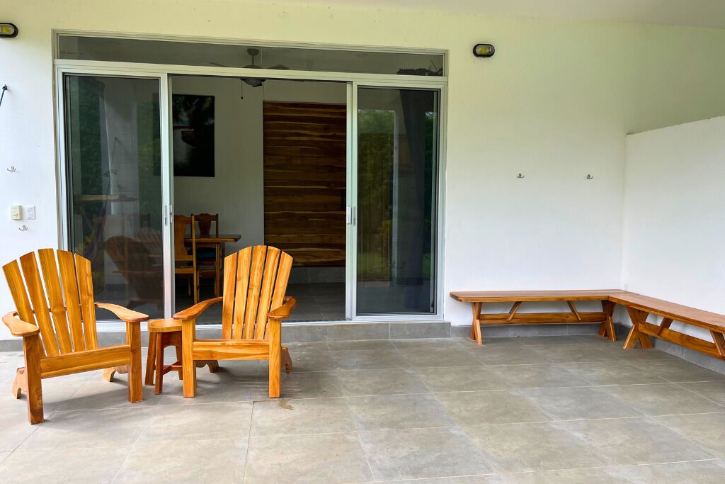 Un patio con sillas y un banco de madera, una puerta corrediza de vidrio que da a un espacio interior, y un piso de baldosas.