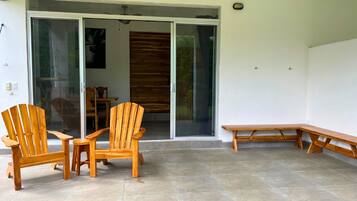 Un patio con sillas y un banco de madera, una puerta corrediza de vidrio que da a un espacio interior, y un piso de baldosas.