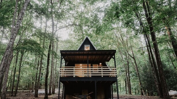 Une cabane à deux étages située dans une forêt, dotée d’une terrasse en bois et d’un porche couvert.