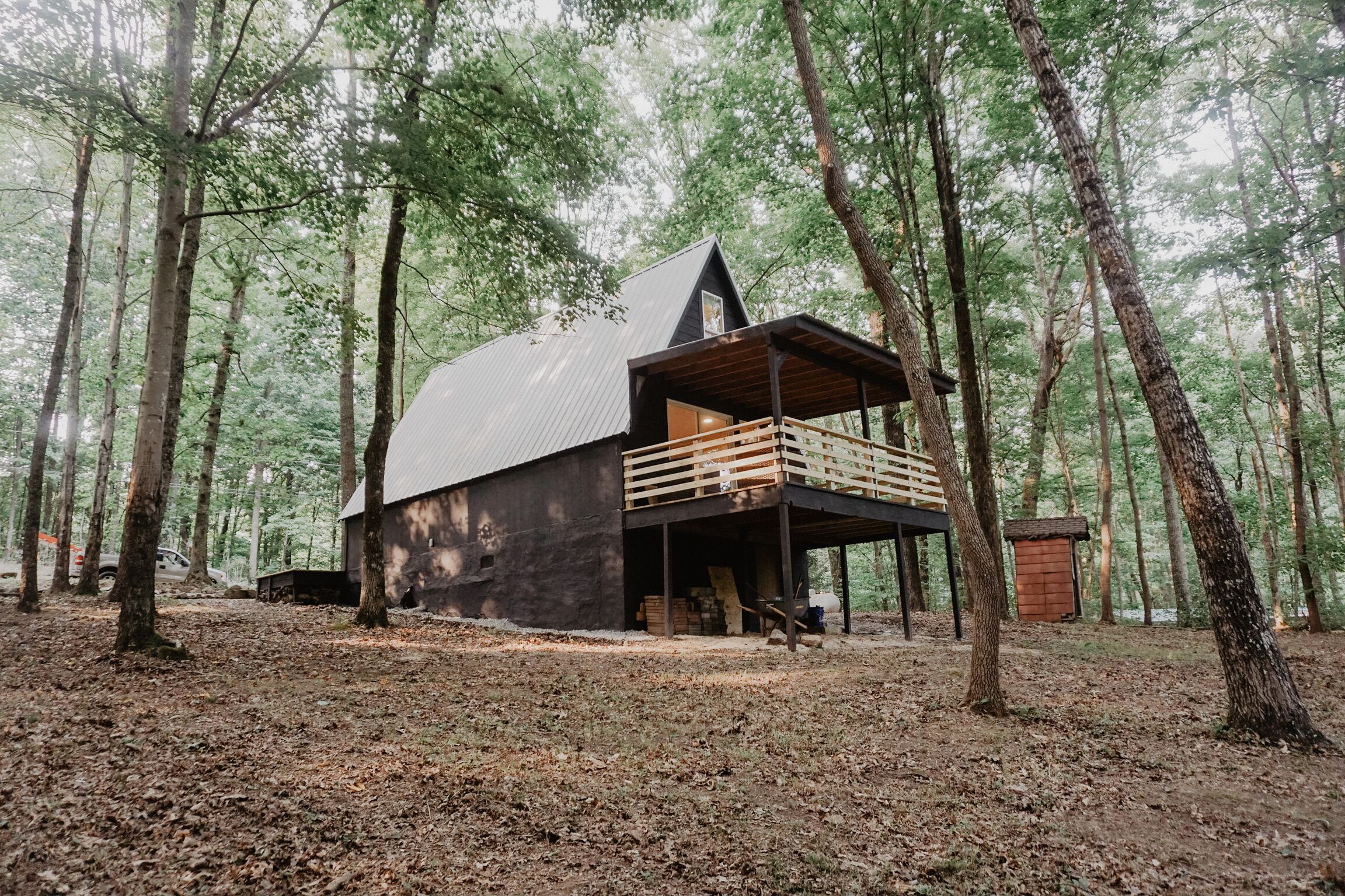 A cabin in a forest with a metal roof, wooden deck, and a small shed nearby.